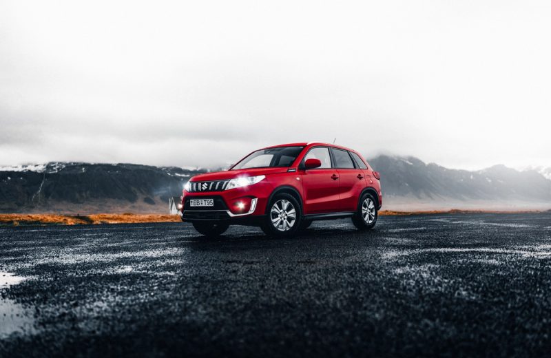 Red Suzuki Vitara 4x4 SUV parked on wet volcanic road in Iceland with mountains behind